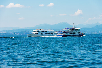 Two ferry boats in motion on Lake Garda (Lago di Garda), in front of the small village of Bardolino, Verona province, Veneto, Italy, Europe. On the horizon the coast of Lombardy with the mountains.