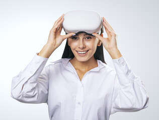 Virtual reality concept: smiled young woman in light shirt with VR glasses on her head on light grey background, close up