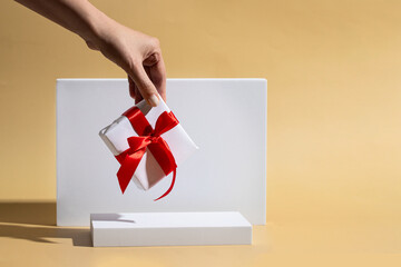 Hand holding white gift box with red bow against the white podiums on beige background.Gifting...