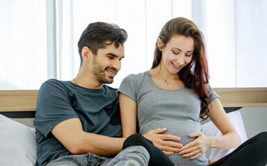 Caucasian husband lies down on belly of pregnant wife on bed at home to embrace unborn baby and feel kicking growth while mom tenderly touching stomach with care