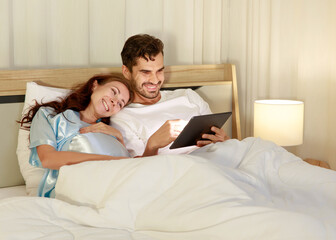 Fototapeta premium Happy pregnant wife holding belly and relax on bed beside beloved husband and enjoy watching entertainment media on tablet together with smile and laugh. Parents expecting joyful life for child birth.