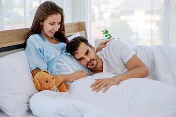 Happy Caucasian husband tenderly lies down on belly and embrace lovely pregnant wife who smile on bed