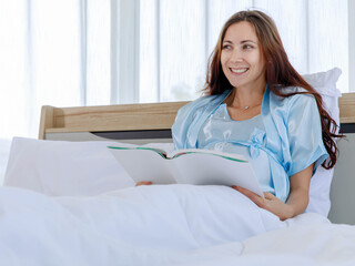 Beautiful pregnant woman on pajamas resting on bed with smile and happy to read book for prenatal care of maternity