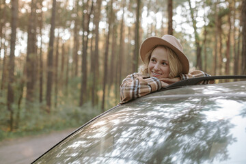Young woman leaning on a car and looking at the forest. Road trip.