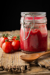 Close-up of homemade ketchup bottle on rustic wooden table with spoon and cherry tomatoes, black background, vertical