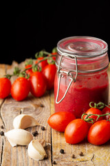 Top view of homemade ketchup jar on rustic wooden table with pepper, garlic and cherry tomatoes, selective focus, black background, vertical