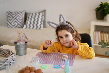 Portrait girl with bunny ears in yellow sweater looks at the camera painting Easter eggs on the table of her home