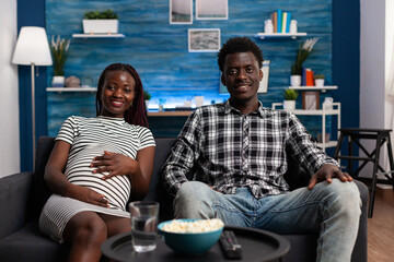 Husband and pregnant wife sitting on sofa and watching television show while having a leisure time together. Happy african american couple sitting on couch in living room expecting newborn.
