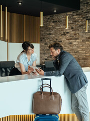 Businessman Using Digital Tablet to Checking In at Hotel. 
Businessman with luggage standing at hotel reception and filling in registration forms on digital tablet with assistance of female concierge. - obrazy, fototapety, plakaty