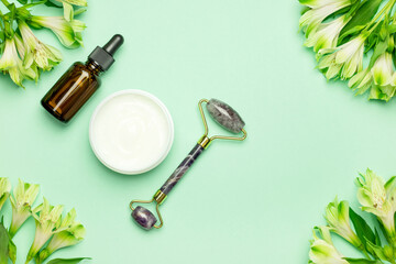 Face roller with cosmetic serum and face cream on a green background with flowers. Facial massage kit for lifting massage.