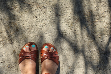 Women feet with blue turquoise pedicure in brown greek leather sandals on grungy cement floor road with tree leaves shadows in sunlight.