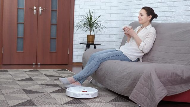 Easy floor cleaning. A view of a round automatic vacuum cleaner on the carpet by a smiling woman with a mag of tea. A concept of easy and lazy housekeeping.
