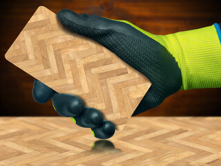Close-up of a hand with protective work gloves holding a herringbone wood parquet sample, on top of a similar floor, with shadows and reflections.