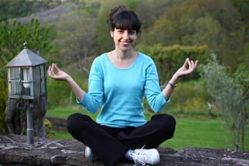 Fototapeta premium Beautiful brunette woman with a blue t-shirt practicing yoga with her legs crossed on a wall next to a lantern in the green field of Navarra while she smiles and practices the Lotus position