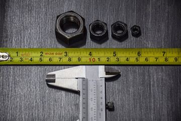 Yellow measuring tape measures the size of a steel plate, textured gray, black pattern. Vernier caliper head below. Choose high-precision measurement. Nuts, screws, small and large arrangement..