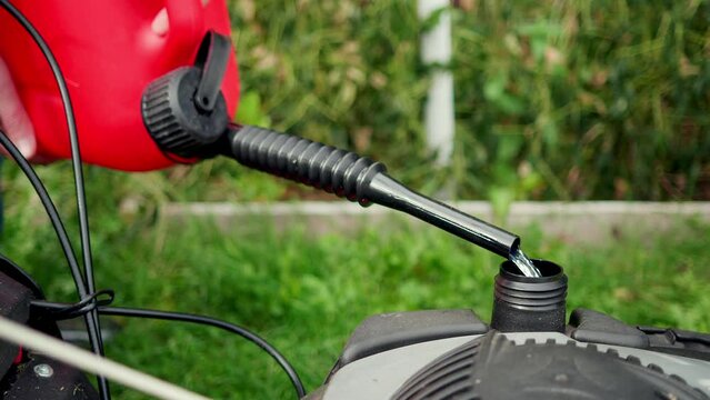 In summer, a gardener pours gasoline from a red plastic canister into the tank of a lawn mower on the street