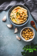 vista superior de un bol con hummus, un bol con garbanzos, dientes de ajo y una cuchara de madera con pimentón, sobre fondo azulado y con espacio en blanco