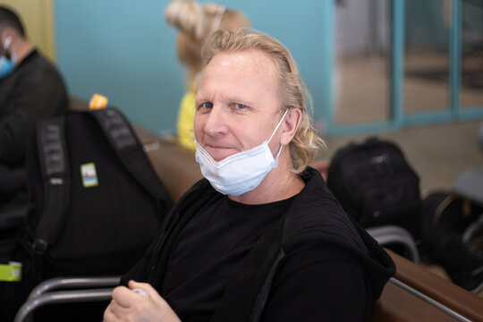 A Blond European Man Sits At The Airport In A Protective Mask Lowered To His Chin And Smiles. Prevention Of Covid In Crowded Places.