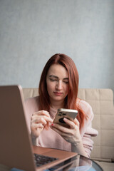 Redhead young woman sitting on sofa with laptop and smartphone.