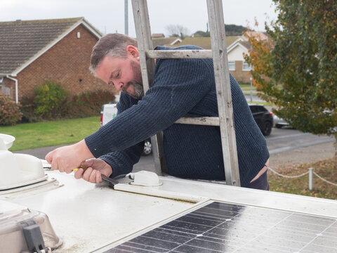 A man can be seen working from a ladder to remove plastic ducting from the roof top of a motorhome recreational vehicle.Solar panel visible.Viewed at roof top height.
