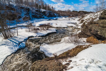 A spring landscape with a river, rocks, ice, snow, trees, a ruined brick building and a blue sky with white clouds