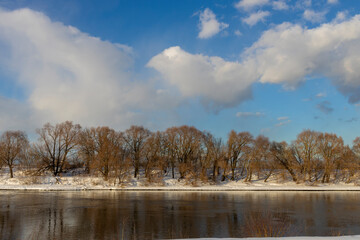 Obraz premium View of the river on a sunny winter day. Trees on the banks of the river illuminated by bright sunlight.