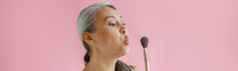 Funny middle aged Asian lady wrapped with white towel blows cosmetic brush posing on pink...