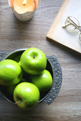 Bowl of green apples, open book, reading glasses, lit candle and flowers on the table. Top view.