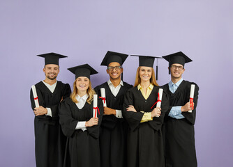Portrait of group of happy multiracial university graduates with diplomas in their hands on purple background. Students dressed in mortarboards and in bachelor mantles stand in row and look at camera.