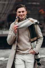  Young man wearing coat walking in the street. Young bearded guy with modern hairstyle in urban background