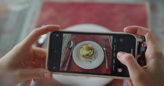 a girl at breakfast photographs a sandwich with a poached egg on her phone. white hands hold the phone and take off the food. dish through the phone screen. close-up