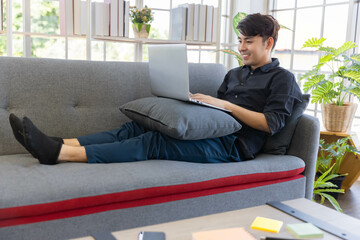 An Asian young business man enjoy work from home by his computer notebook on his sofa in living room at home
