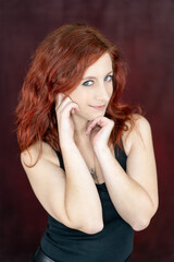 attractive red-haired girl smiling while looking at the camera from the corner of her eye and placing her hands on her face with a red studio backdrop