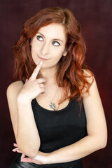Obraz premium beautiful red-haired woman thinking with one finger of her hand touching her chin against a red studio backdrop