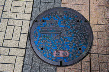 Beautiful manhole cover of a well, a water pipe with a picture of a temple and sakura, on the sidewalk, paving slabs, Tokyo, Japan.