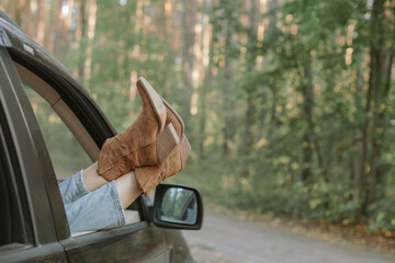 Female legs in trendy boots sticking out of car window in the forest. Road trip.