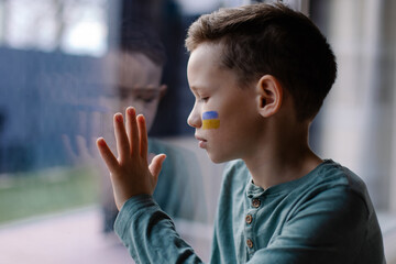 the child sits at the window with the flag of ukraine. State support in war. United Europe against Russia. Hope for the world. Anxiety and anticipation. Children's tears. Save Ukraine