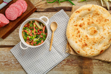 Jewish cuisine bean soup cholit in a tureen on a gray napkin next to a sausage and a flatbread and a wooden spoon.