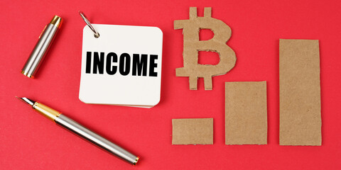 On a red surface lie the bitcoin symbol, a pen and a notepad with the inscription - INCOME
