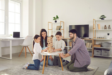 Happy family with kids playing board games at home. Smiling mom, dad and children sitting on the floor around a low table in a cozy living room and pulling wooden blocks from the tower