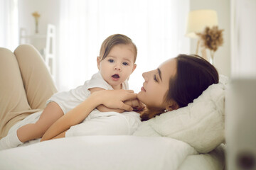 Happy mother together with her child cuddling on comfy bed at home. Young mommy and adorable baby in comfortable white cotton bodysuit lying on bed in the morning or afternoon. Family and care concept