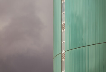 View of the green facade of a tall modern building with a vertical row of narrow windows and the cloudy sky on the left