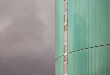 View of the green facade of a tall modern building with a vertical row of narrow windows and the cloudy sky on the left