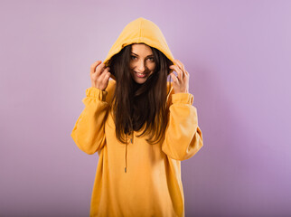 Naklejka premium Relaxing thinking smiling woman posing in fashion yellow hoodie with hood on the head on purple bright background with empty copy space.