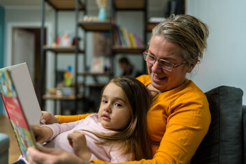 Loving grandmother teaching granddaughter holding book sitting on sofa, grandma baby sitter...