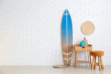 Table with vases, pouf and surfboard near white brick wall in room interior