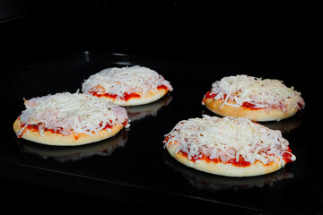 Four uncooked mini pizzas with ham, cheese and sause on tray in electric oven, black background. Italian cuisine, homemade bakery, fast food, semi-finished products and raw concept - obrazy, fototapety, plakaty