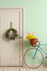 Bicycle with tulips and bunny ears near wooden door with Easter wreath in room