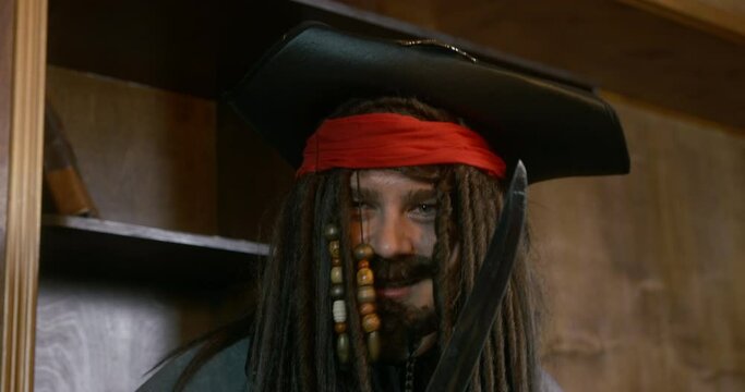Crazy pirate captain with dark long braids in cocked hat swings with saber against wooden racks IN ship cabin close view
