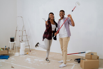 Two friends are renovating dormitory room, they are working together painting walls holding paint rollers in hands, buckets ladder with paint next to them smiling woman is standing next to man